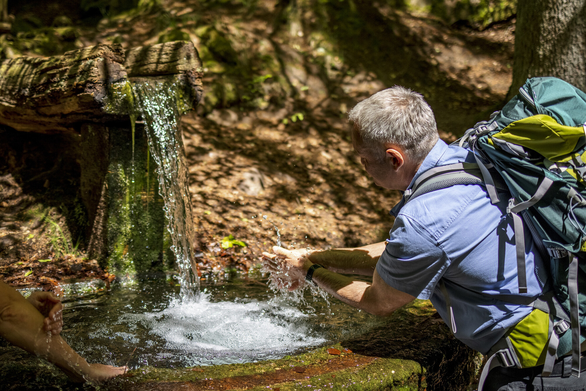 Wanderer an Quelle Ein Wanderer mit Rucksack hält seine Hände unter einen kleinen Wasserfall.