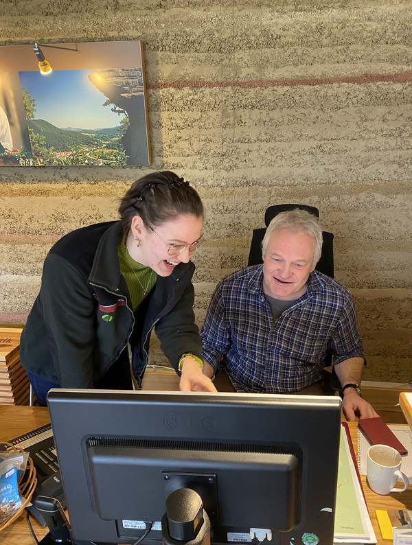 Eine junge Frau hilft einem älteren Herrn am PC, beide lachen.