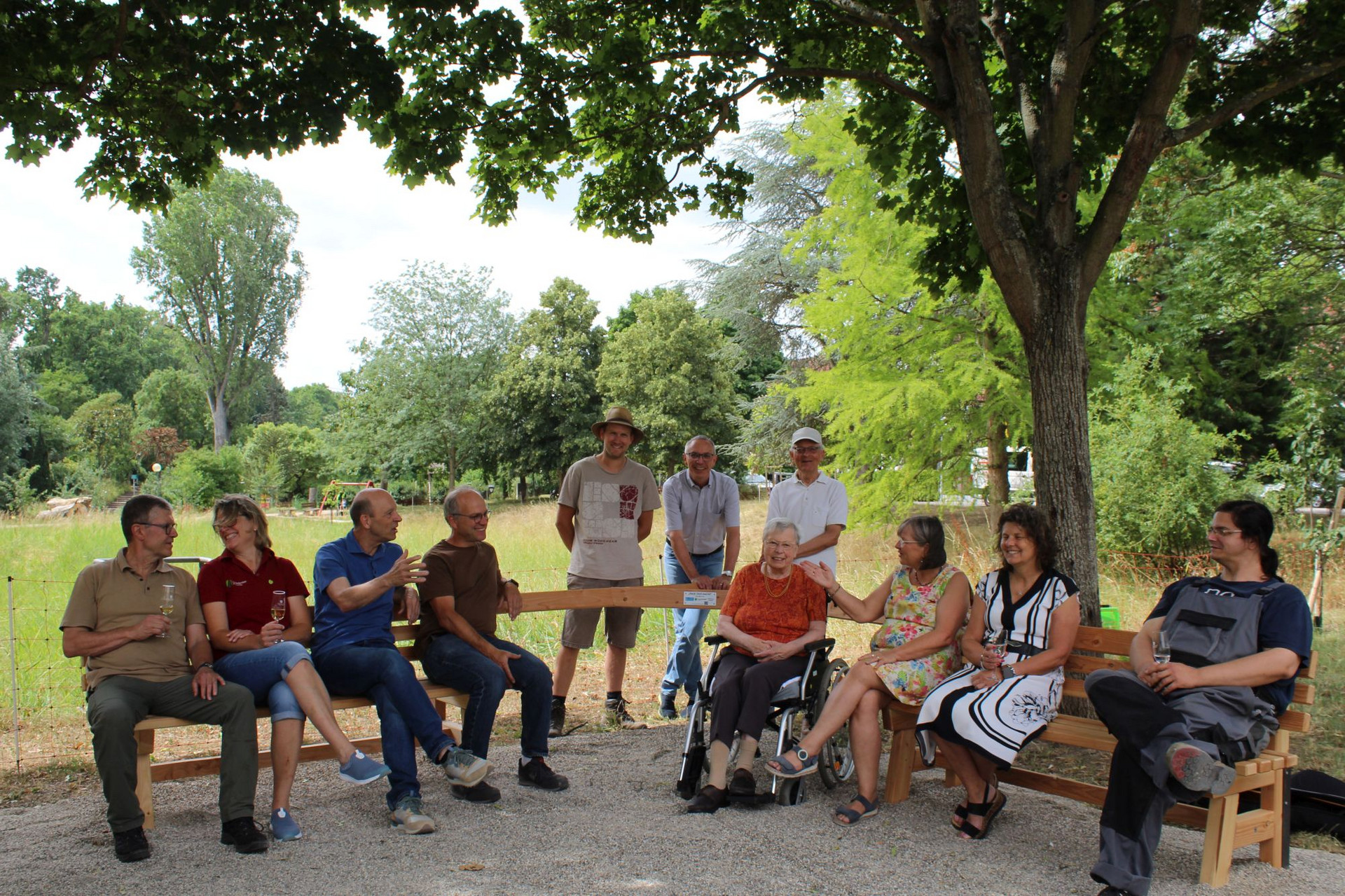 Begegnungsbank Deidesheim Die Begegnungsbank steht unter Bäumen, im Hintergrund Wiese und Büsche. Viele Leute sitzen auf der Bank und unterhalten sich.