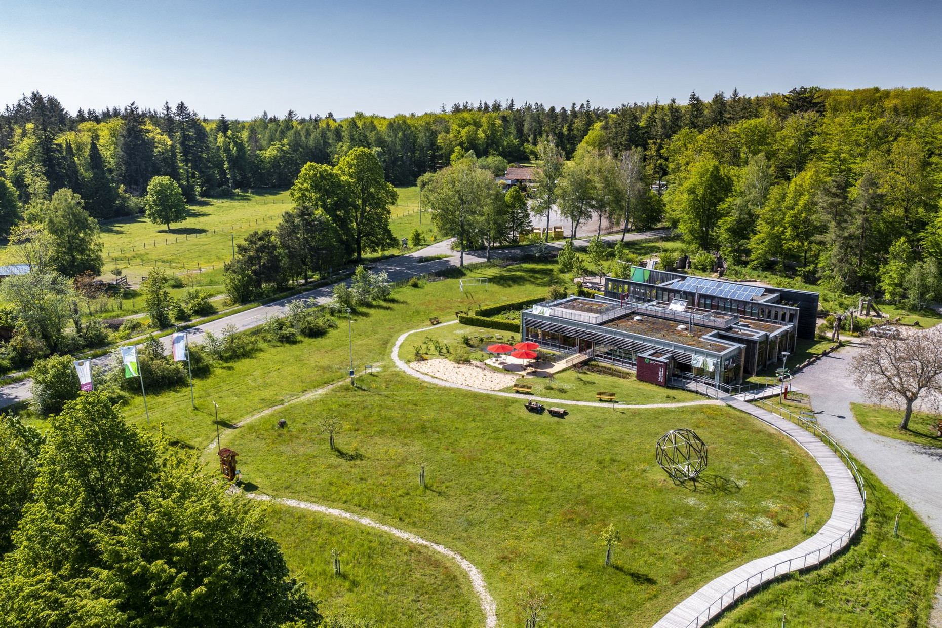Das Gebäude und sein Umfeld (Wiese, Terrasse, Parkplatz) im Sonnenschein.