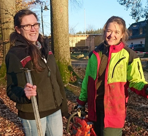 Das Bild zeigt zwei junge Frauen im Wald, die mit Arbeitskleidung Motorsäge und anderem Werkzeug ausgerüstet sind.