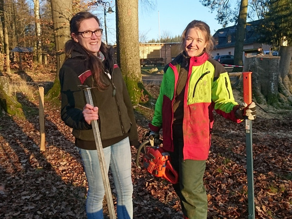 Das Bild zeigt zwei junge Frauen im Wald, die mit Arbeitskleidung Motorsäge und anderem Werkzeug ausgerüstet sind.