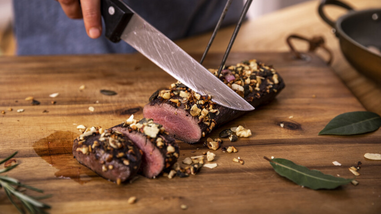 Rehrücken Das Bild zeigt eine Person, die einen fertig zubereiteten Rehrücken auf einem Holzbrett in kleine Portionen schneidet