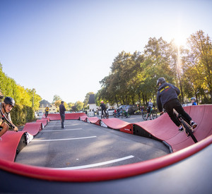 Ein Pumptrack sorgt für Action! zwei Menschen auf Fahrrädern fahren über einen roten geschwungenen Parcours