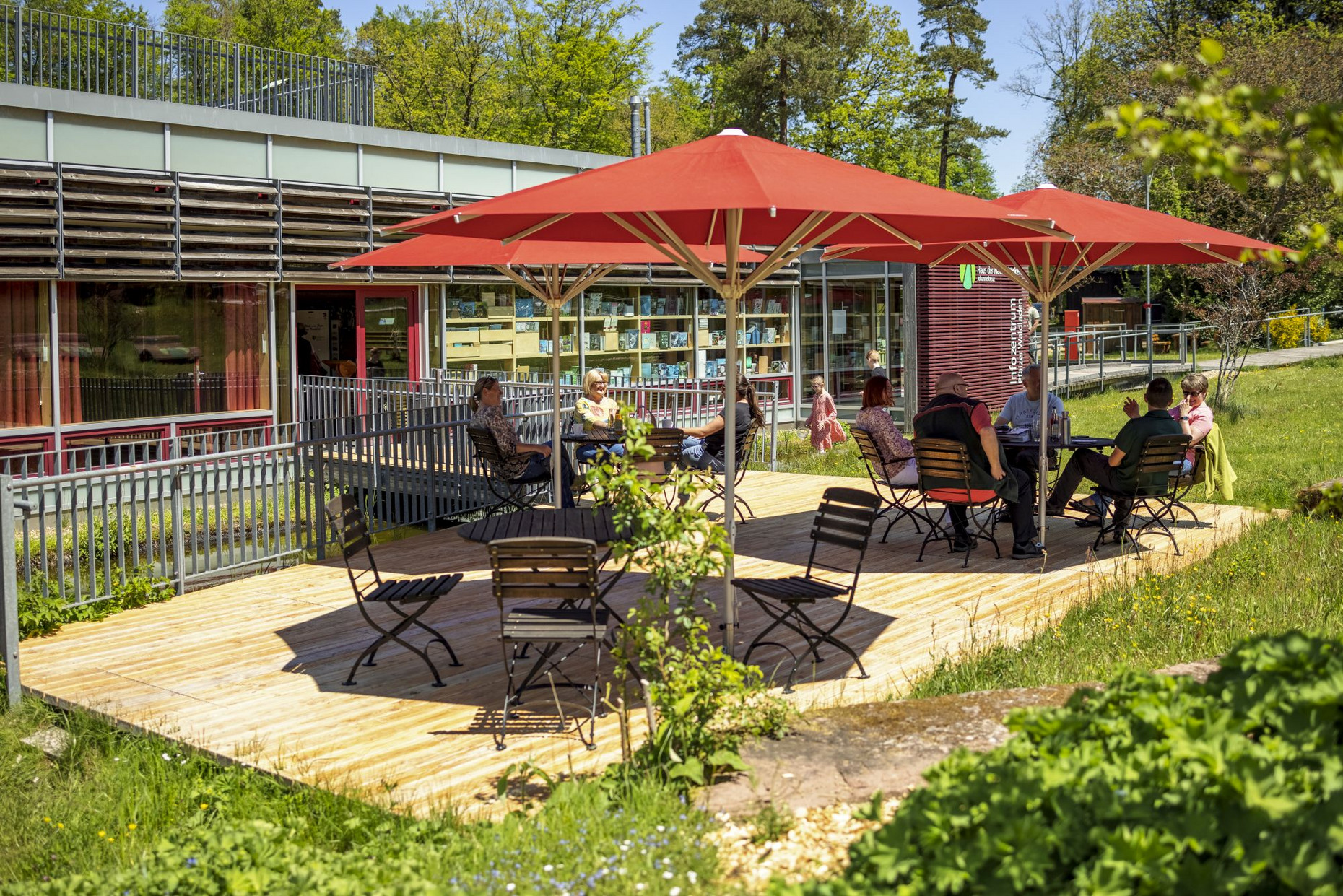 Das Bild zeigt die Teichterrasse des HdN mit roten Sonnenschirmen und mehreren Personen.