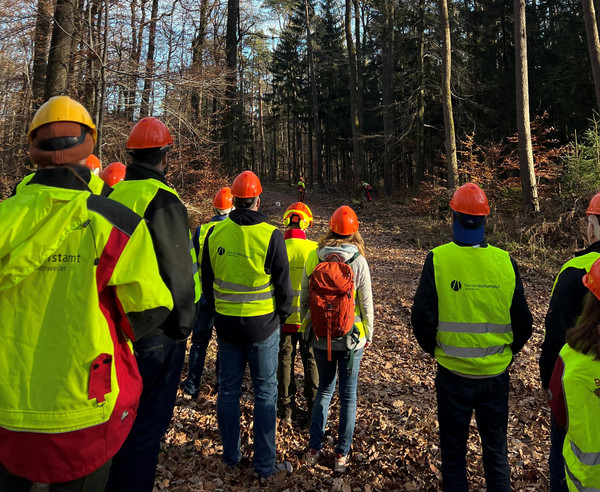 Das Bild zeigt eine Gruppe Menschen mit Warnwesten und Helmen im Wald.